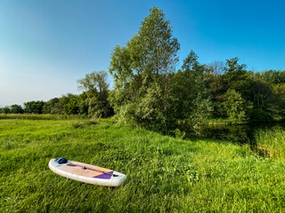 A brightly colored paddle board is gently resting in the lush, green grass, situated near a serene river that flows gracefully through the beautiful, natural landscape in the vicinity