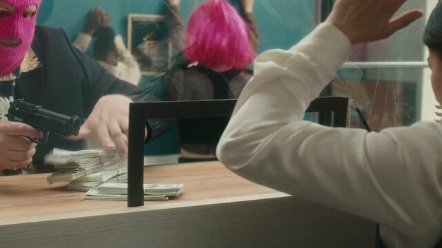 Medium shot of female bank cashier at tellers counter giving out money in cash to masked male robber pointing gun at woman then calling police with shaking hands