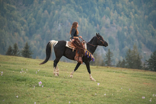 Confident young woman, a cowgirl with spirit of freedom and adventure riding a majestic black horse, through breathtaking mountain terrain