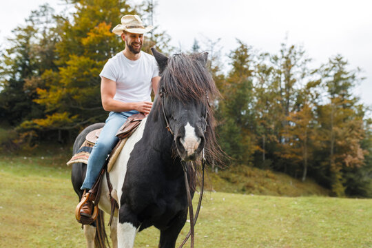 Young man with a cowboy hat rides a black and white horse in a green field. A woman watches in the background on a cloudy day near trees.