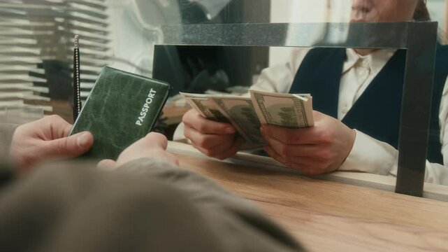 Close up on hands of female bank worker counting dollar bills assisting male customer with money exchange while client holding passport at tellers counter