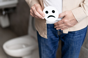 Senior man holding sad face emoticon near groin in bathroom: prostate problems concept