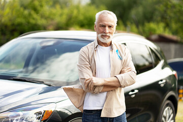 Senior man wearing blue ribbon crossing arms in front of car
