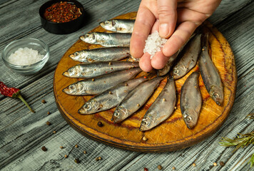 A hand sprinkles coarse salt over fresh small fish arranged neatly on a wooden cutting board....