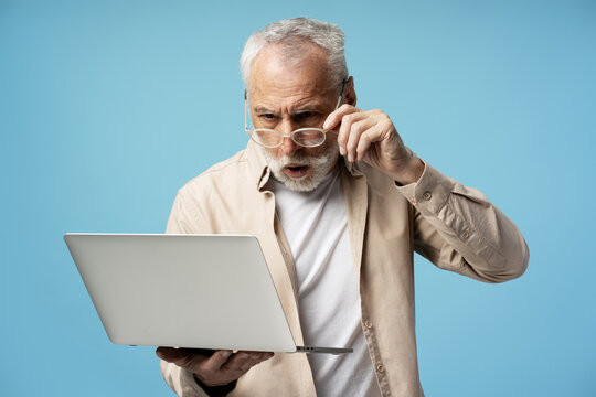 Shocked senior man looking at laptop and adjusting glasses on blue background