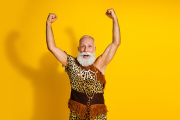 Photo of excited old man in caveman costume raise fists up winning wear animal fur isolated yellow color background