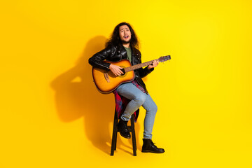 Young musician in a stylish leather jacket playing acoustic guitar on a stool against a vibrant yellow background