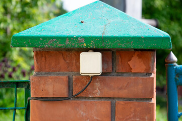 A square electrical box is mounted on a brick fence post topped with a green slanted roof,...