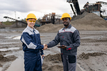 Male surveying engineers in sand quarry shaking hands amidst ore mining equipment, Portrait worker...