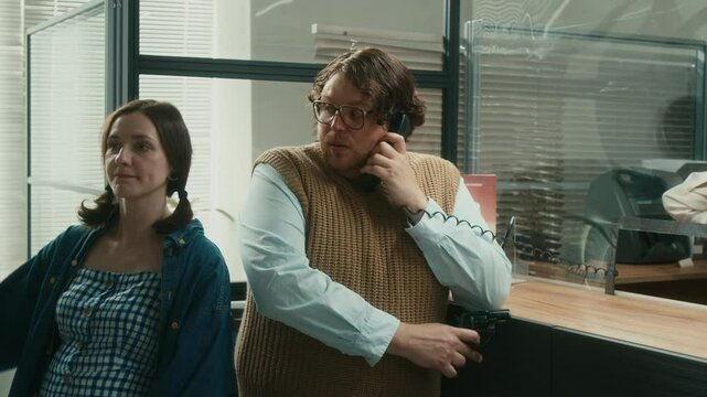 Medium full shot of unprofessional male criminal on phone call speaking to police during hostage negotiation while robbing bank with pregnant female partner holding gun