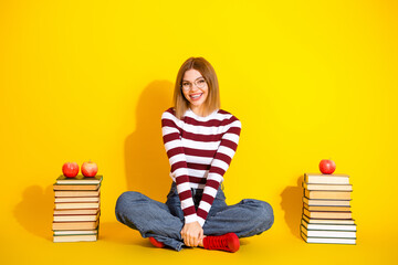 Portrait of a cheerful young woman sitting surrounded by book, featuring a bright yellow background...