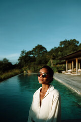 Fototapeta premium Chic woman sunbathing by turquoise pool in stylish sunglasses and white dress