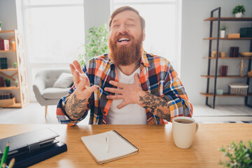 Bearded man in a checkered shirt laughing heartily at his desk while enjoying coffee, evoking warm...