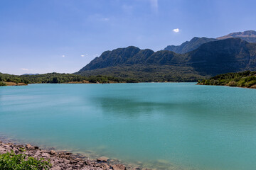 Tranquil waters of Castel San Vincenzo lake under a clear blue sky, surrounded by forested hills and the distant village
