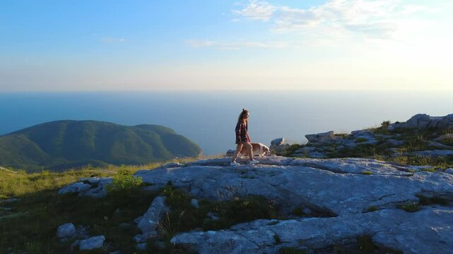 Young Woman Hiking with Dog on Rocky Mountain Trail Overlooking Ocean at Sunset in Summer