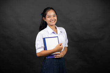 Obraz premium An Asian high school girl poses in a studio background, holding a notebook from her school, imagining her bright future at a leading university where she plans to study science.