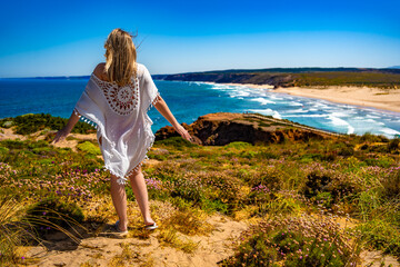 Beautiful female model posing backwards in flowing dress standing at viewpoint on sandy beach...