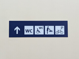 Informational sign on a light wall directing to public toilets, accessible restroom, baby changing station, and bicycle parking — practical facilities in a public building or transportation hub.