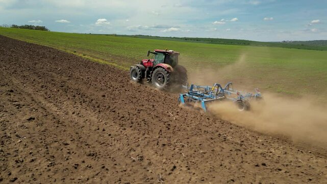 Aerial tracking footage of tractor pulling disc harrows attachment in a rural farm field. Combine harvester plowing earth to prepare for crop planting. Tilling soil by industrial machine in rural farm