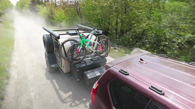 A family enjoys a camping trip as they travel down a dusty road in a teardrop trailer, bicycles secured on the rear, surrounded by lush green trees.