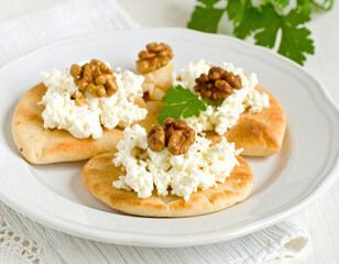 Three pieces of soft, round flatbread are topped with creamy white cheese and crunchy walnuts, garnished with a fresh parsley leaf on a white ceramic plate.