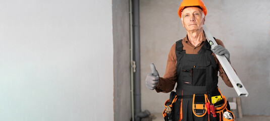 old man builder construction worker with spirit level and tools standing on construction site showing like gesture class raising thumb up