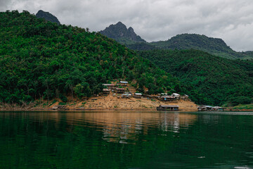 View of a rural community's simple dwellings built along a water body, with dense tropical forests covering the towering hills in the background.