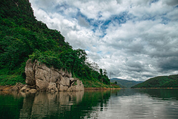Panoramic view of a tranquil lake reflecting an overcast sky, with dense tropical forest clinging...