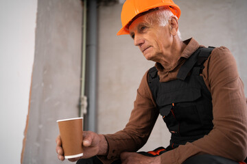 Elderly Builder in working uniform drinking coffee sitting at construction site. building and industry