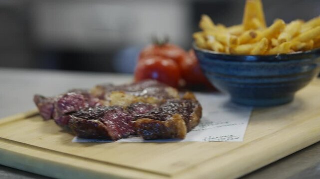A person is cutting a piece of meat on a wooden cutting board. The meat is on a paper towel. A bowl of fries is sitting next to the meat