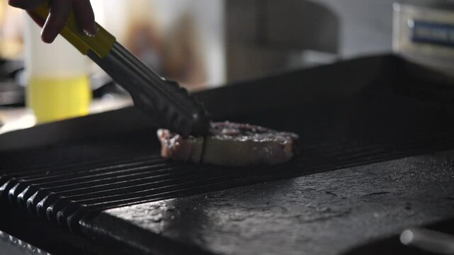 A person is cooking meat on a grill. The meat is being flipped with tongs. There is a bottle of oil on the counter