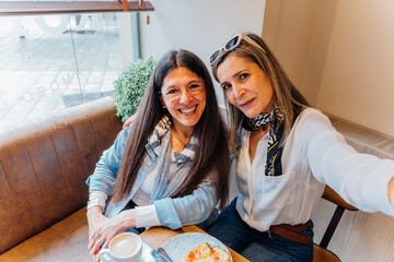 Mature female friends smiling, taking selfie while sharing coffee and treats at intimate cafe...