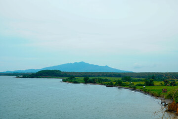 a calm sea view with agricultural rice fields and a backdrop of cloud-covered mountains.