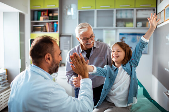 Happy young girl high-fiving her doctor in a medical office with her smiling grandfather standing beside her