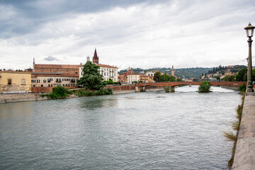 A scenic view of Verona along the Adige River, with historic buildings and lush hills in the background on a cloudy day