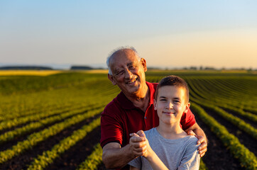 Portrait of senior farmer with his grandson standing in green soybean field and looking at camera.