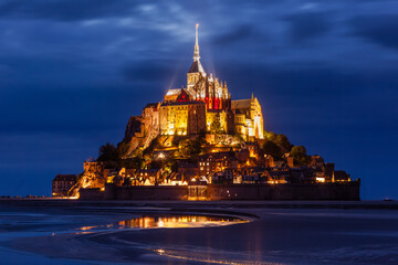 Mont Saint-Michel abbey illuminated at night, under stormy sky, with mirror reflection in calm water, listed as World Heritage by UNESCO, shortly after sunset, Normandy, Manche, France
