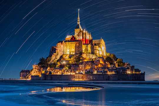 Mont Saint Michel abbey illuminated at night, under circumpolar star trails, with mirror reflection in calm water, summer starry night, Night of the Stars (Nuit des Etoiles), Normandy, Manche, France