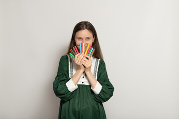 A young woman stands in a studio, holding a collection of vibrant markers close to her face. She...