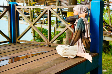 Asian Muslim woman sits cross-legged on the wooden floor, leaning against a pillar while massaging...
