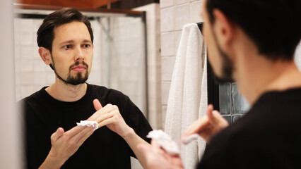 Young man applying shaving foam to cheeks while looking in mirror
