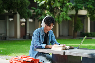 Portrait of a young Asian male college student e-learning using laptop on the bench on campus.
