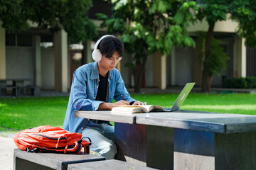 Portrait of a young Asian male college student e-learning using laptop on the bench on campus.