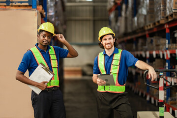 Friendly warehouse staff member smiles brightly, holding the brim of his helmet while standing in the aisle, card box and trolley. Worker in logistic portrait.