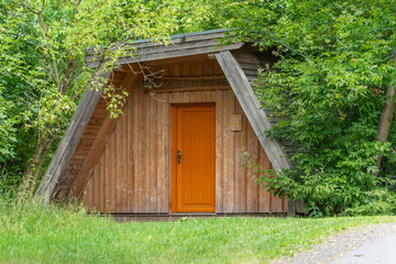 A small wooden hut with an orange door stands surrounded by dense greenery. The construction is...