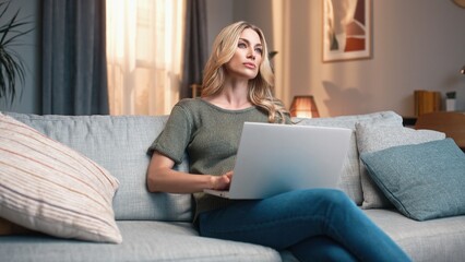 Woman working with laptop sitting on sofa. Female sitting on couch and working distantly. Woman...