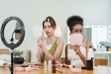 asian female teen and african female teen sit at table applying makeup while looking into mirrors and recording online beauty tutorial together with cosmetics and tools spread across surface