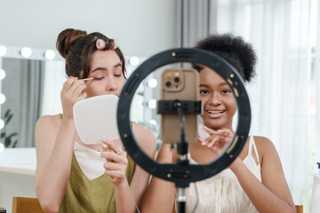 asian female teen applies eyeshadow while african female teen looks at smartphone camera smiling during online beauty tutorial recording with ring light and handheld mirror on desk