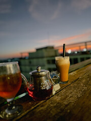 An inviting scene from above shows coffee and tea cups resting on a wooden table, framed by a golden sunset casting warm light over distant hills and sky.
