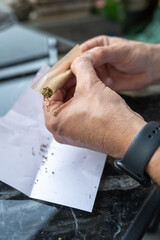 Close-up of a mans hands skillfully rolling a cannabis joint with shredded herb on paper, placed on a sleek marble surface, highlighting a relaxed and precise preparation.